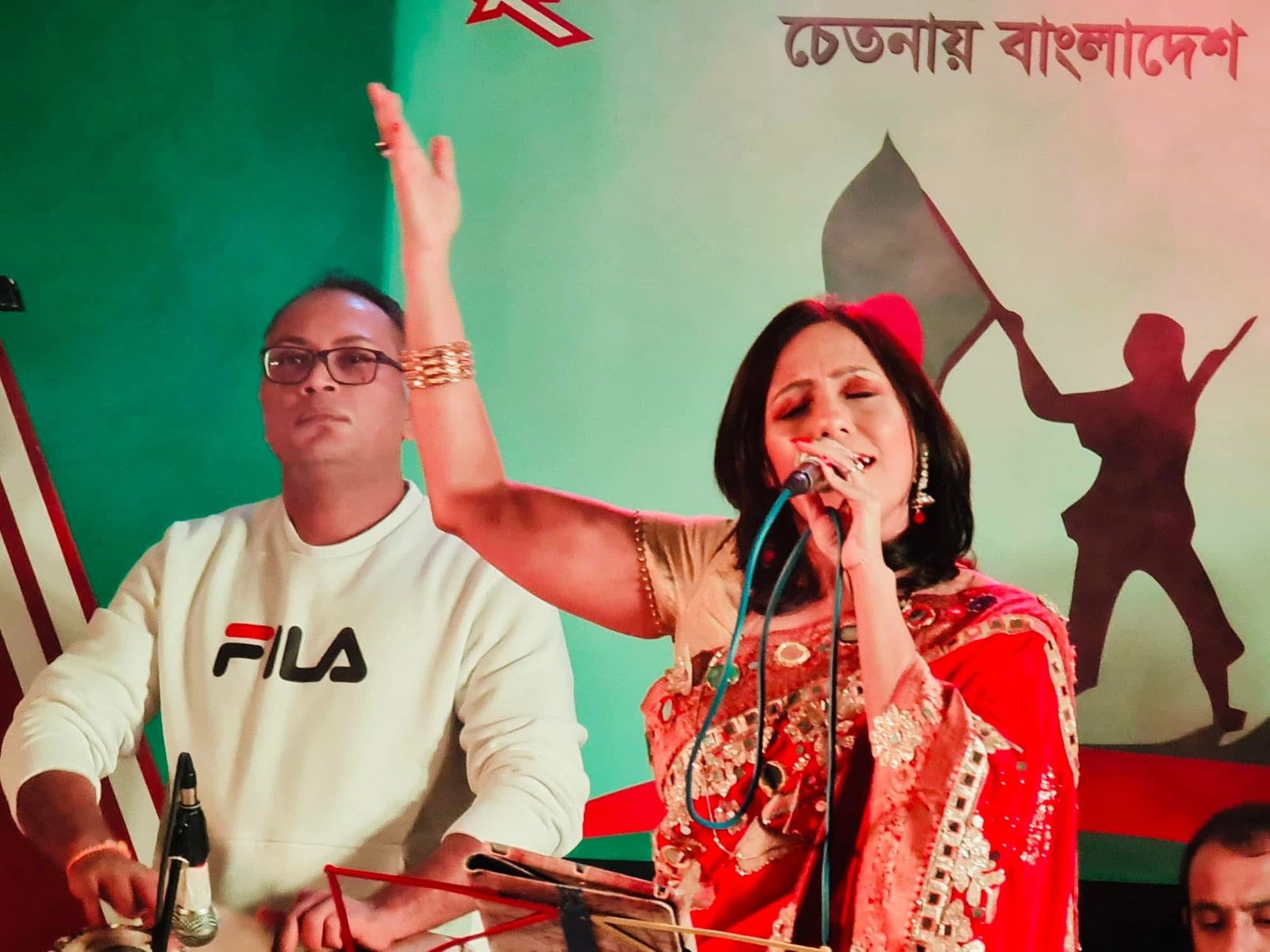 Rowshanara Moni singing with one hand raised in front of a Bengali cultural backdrop
