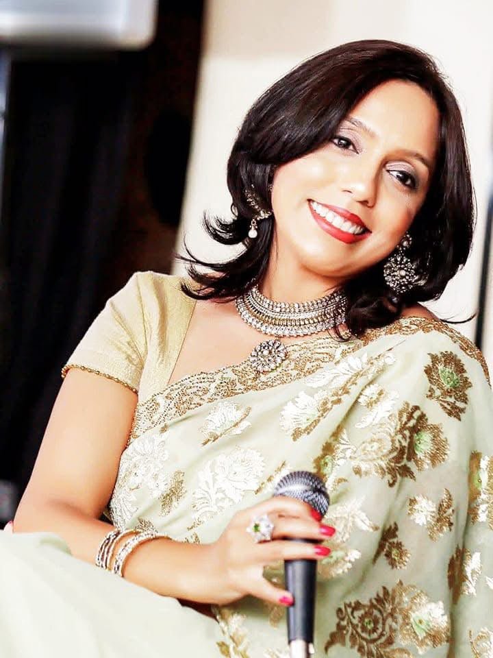 Rowshanara Moni smiling, seated in a pale green and gold embroidered sari, holding a handheld microphone