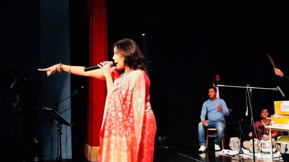 Rowshanara Moni singing into a handheld microphone on a darkened theatre stage in a coral sari, one arm outstretched, with a tabla player and musicians seated behind her