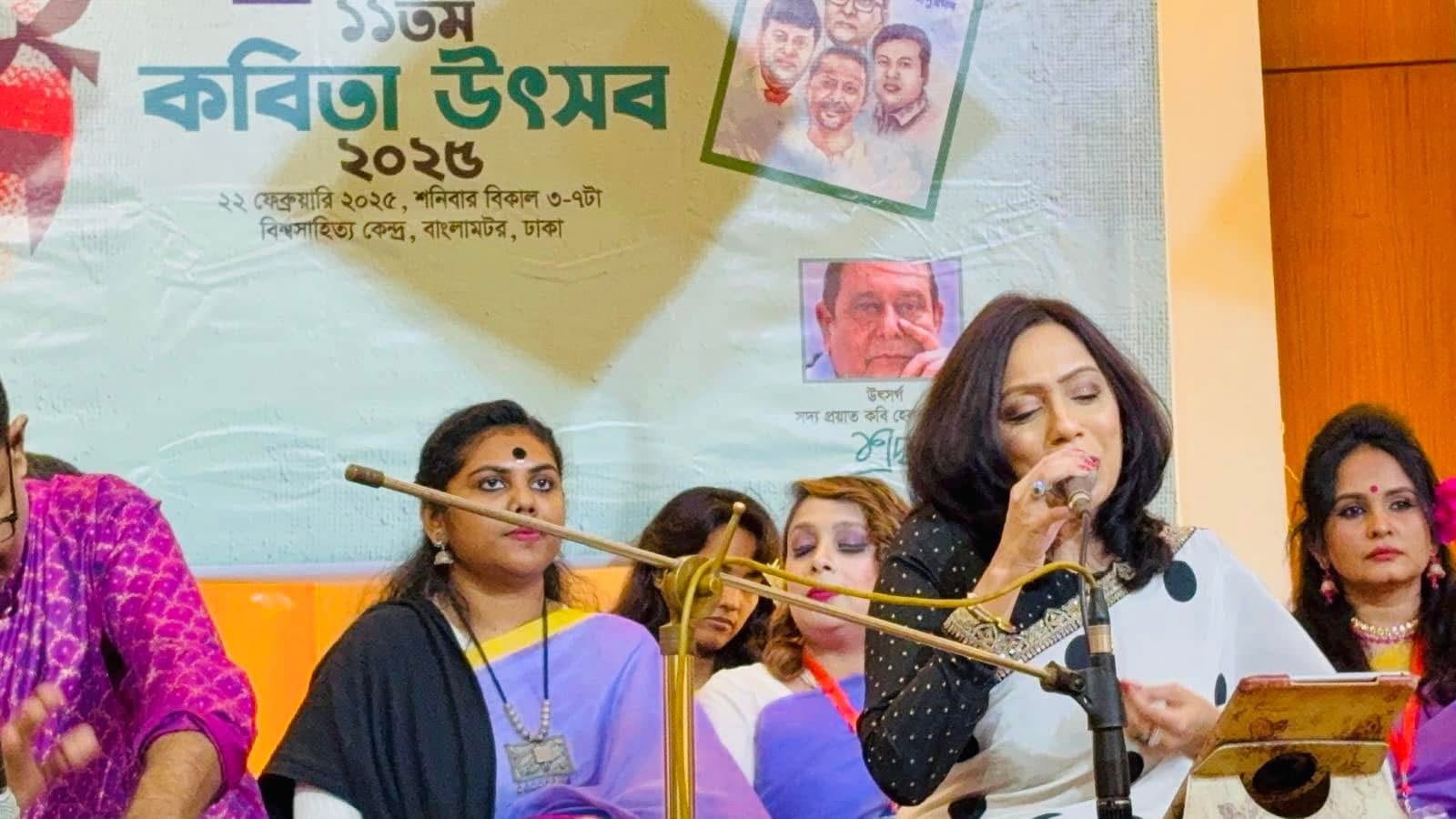 Rowshanara Moni singing into a microphone in a black and white sari, seated among fellow performers beneath the Kobita Utsob 2025 banner in Dhaka