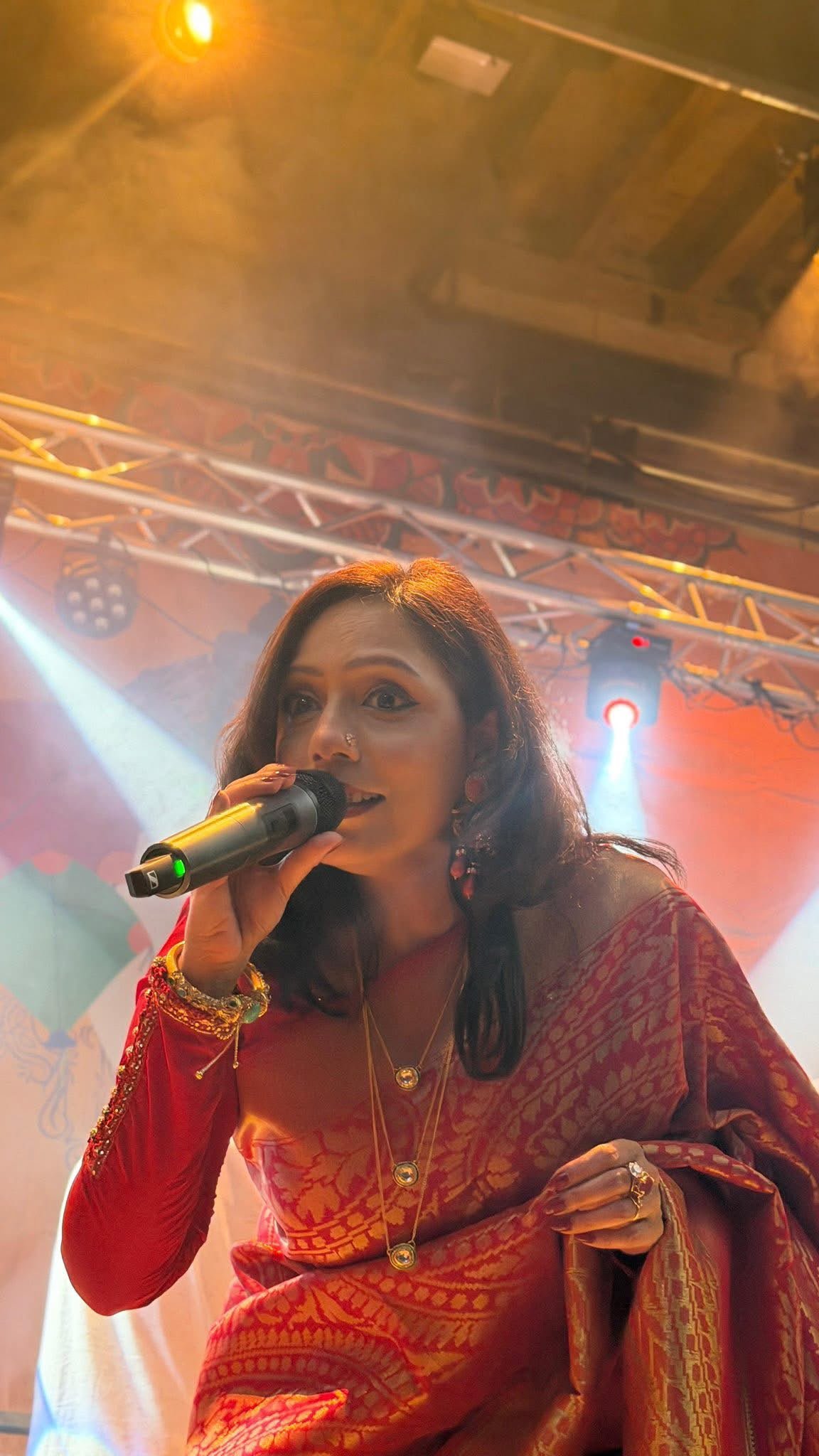 Close portrait of Rowshanara Moni singing into a handheld microphone in a red and gold sari under stage lights