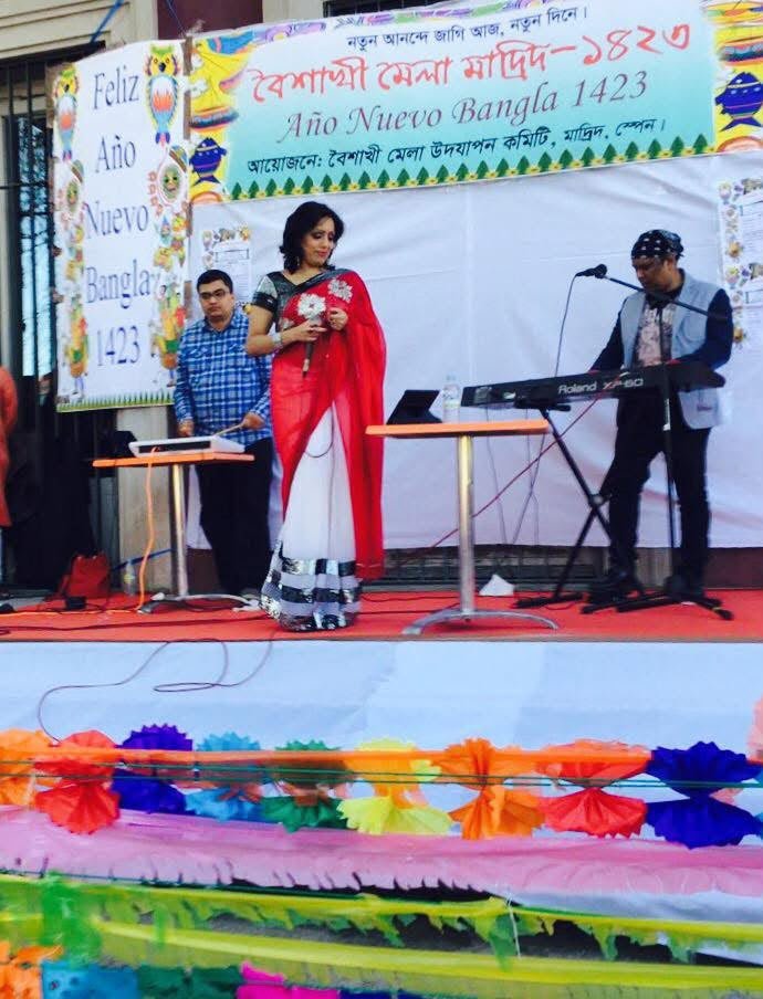 Rowshanara Moni on an outdoor stage in a red and white sari, holding flowers, with a keyboard player beside her