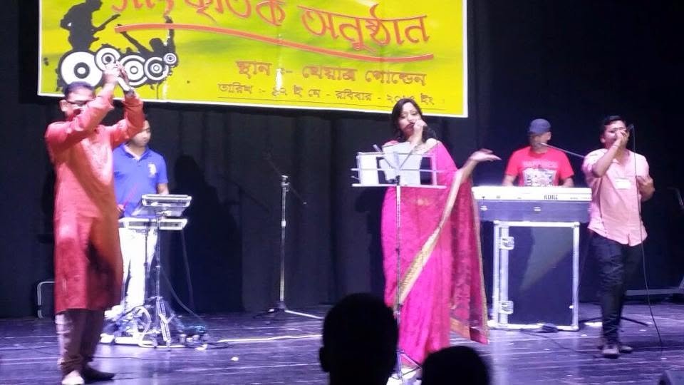 Rowshanara Moni singing centre stage in a pink sari at a microphone stand, with a flautist, keyboard player and fellow vocalists beside her