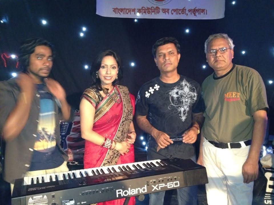 Rowshanara Moni in a red and gold sari standing with three musicians behind a keyboard, string lights in the background