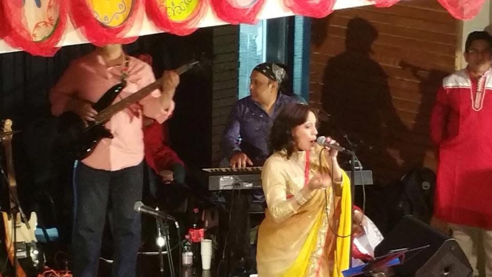 Rowshanara Moni singing with a live band — guitarist and keyboard players — beneath red and white decorations