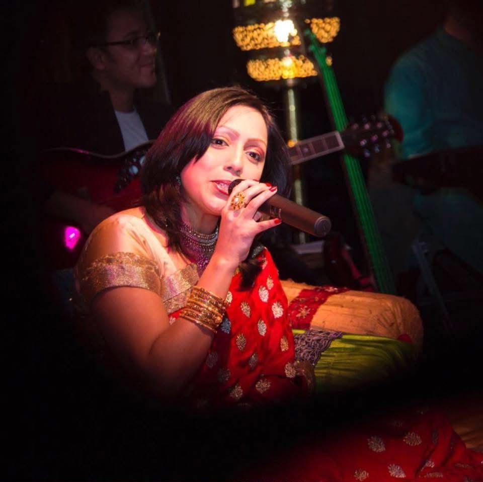 Rowshanara Moni singing into a microphone in a red and gold sari, warm stage lighting behind her