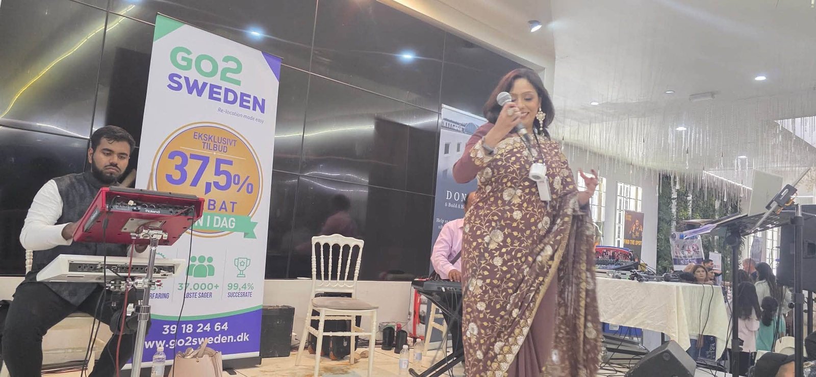 Rowshanara Moni singing into a microphone in an embroidered sari, with a keyboard player to one side of the room