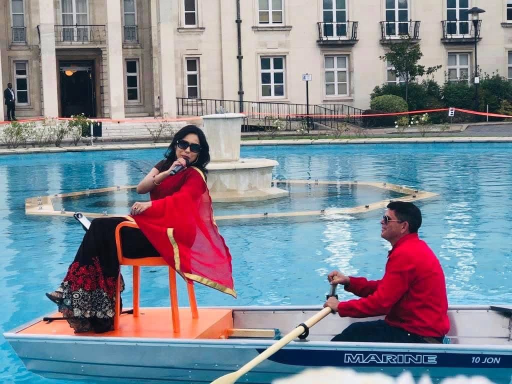 Rowshanara Moni performing in a red sari, seated in a small boat being rowed across an ornamental pool