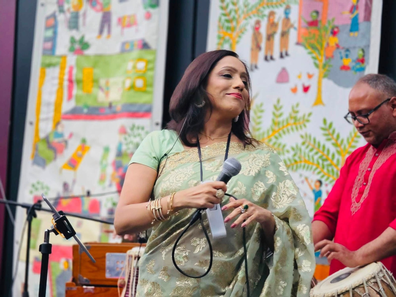 Rowshanara Moni performing on stage in a green and gold saree, holding a microphone