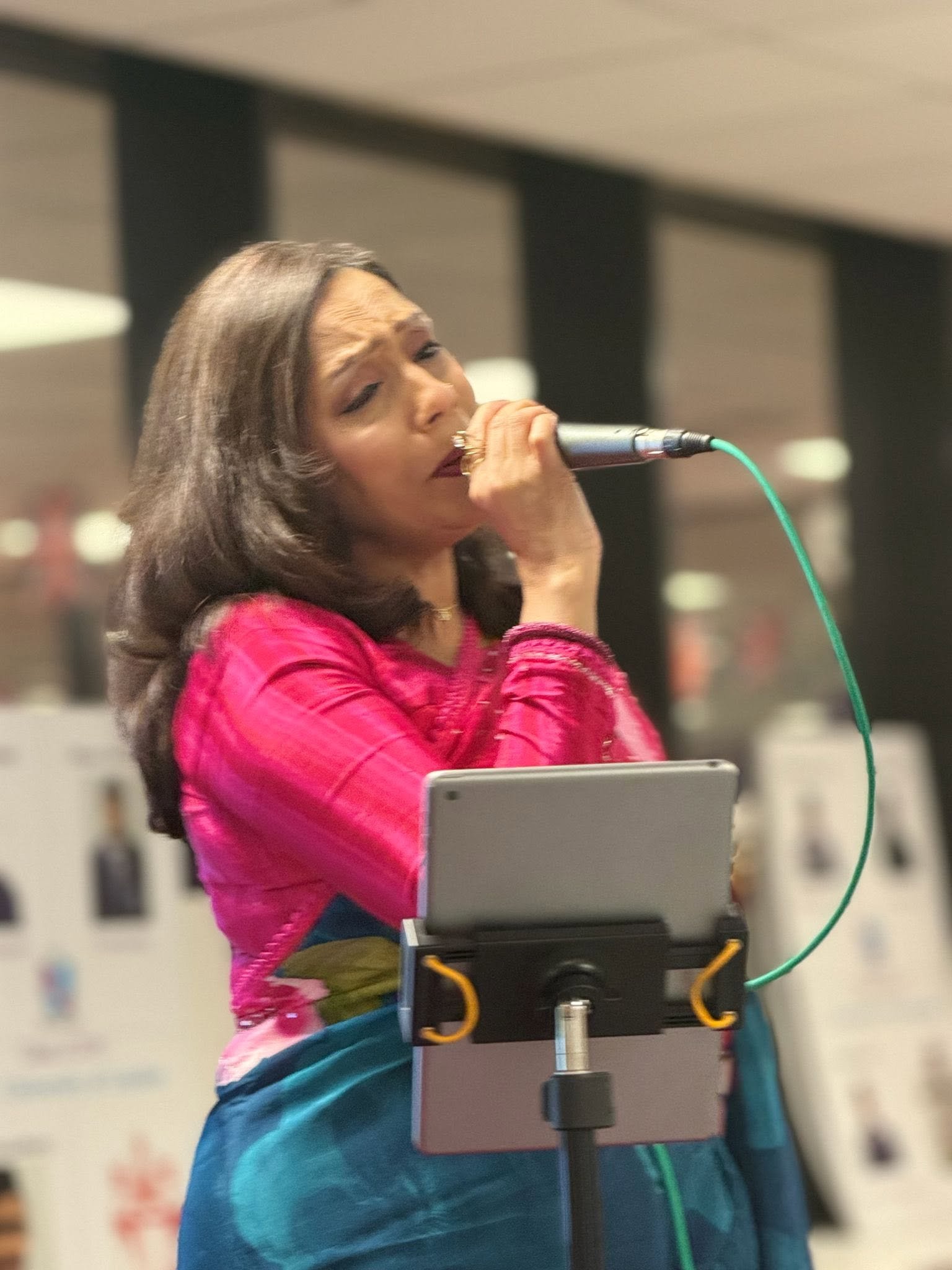 Rowshanara Moni performing, holding a microphone close, wearing a pink and teal sari
