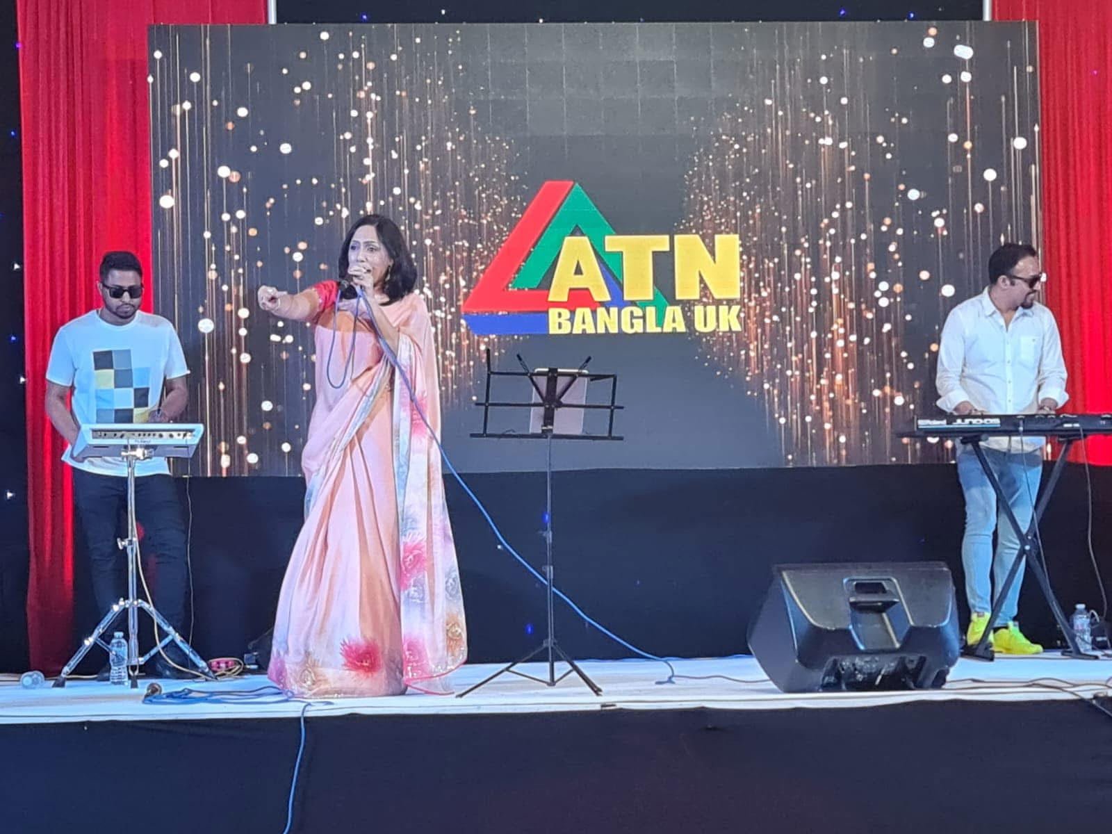 Rowshanara Moni performing on stage in front of a large ATN Bangla UK screen with two keyboard players