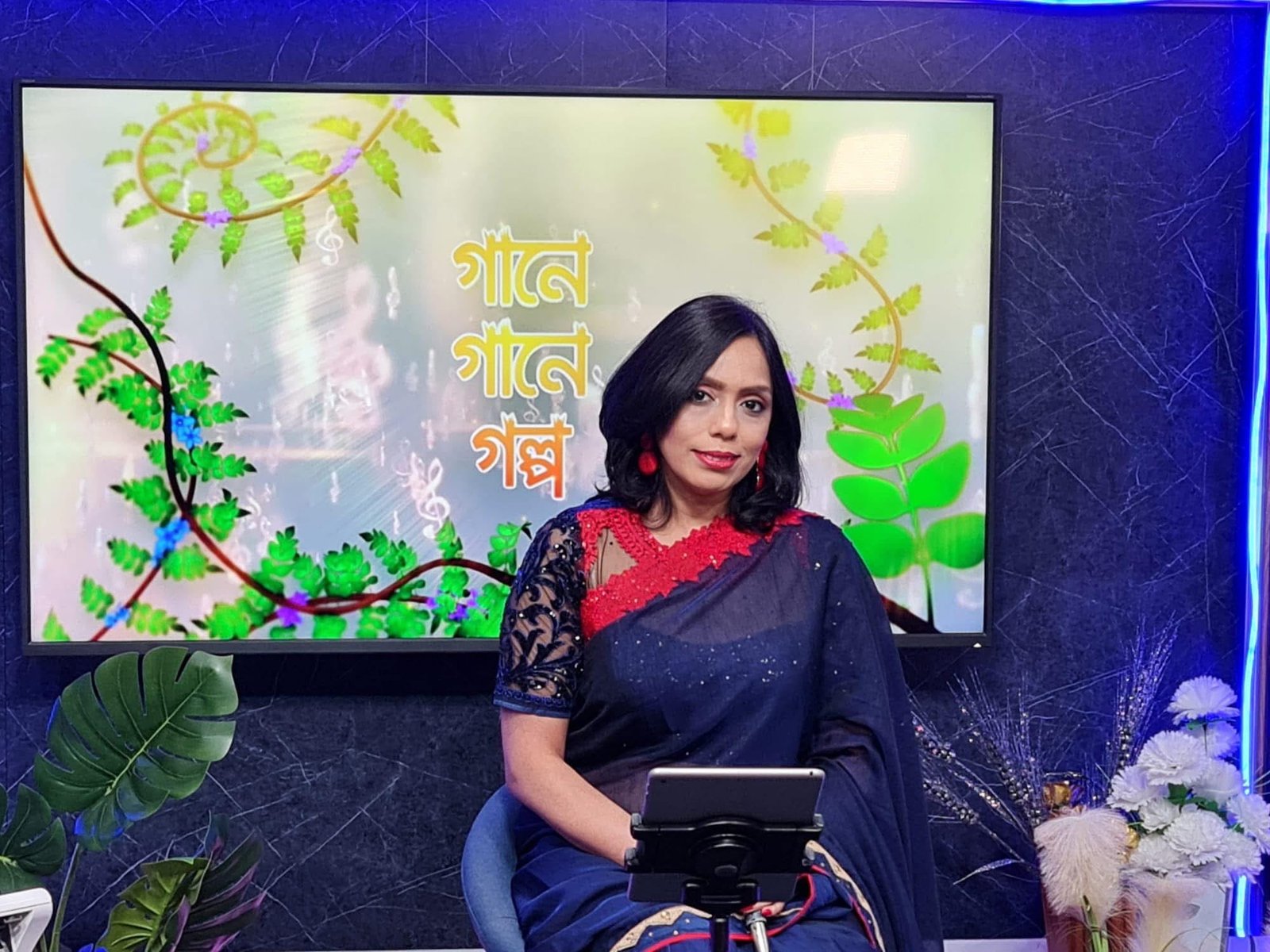 Rowshanara Moni seated in a television studio in front of a Gane Gane Golpo programme screen