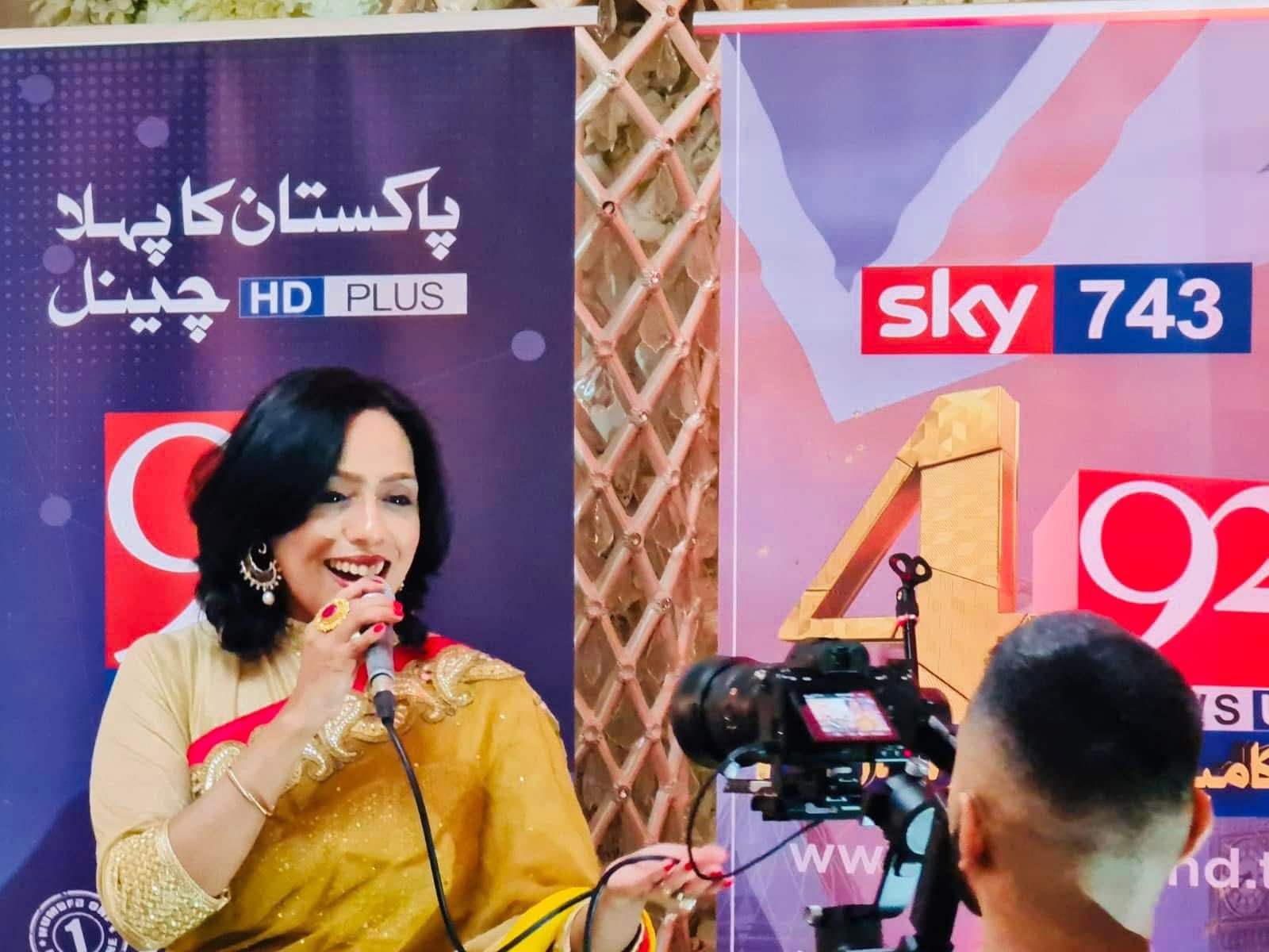 Rowshanara Moni singing into a microphone in front of channel banners while a camera operator films